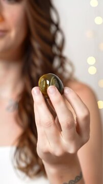 Close-up video: young caucasian woman's hand holds a golden-brown tiger's eye crystal. Rack focus, subtle wrist tattoo, brown wavy hair, soft white top, against luminous bokeh and soft white