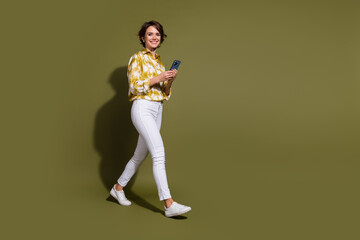 Confident young businesswoman in casual outfit walking with phone against khaki background