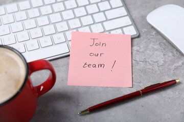 Sticky note with phrase Join Our Team, computer keyboard, mouse, pen and coffee on grey table,...