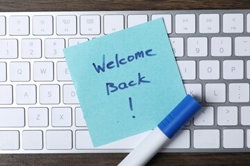 Paper note with phrase Welcome Back, marker and keyboard on table, top view