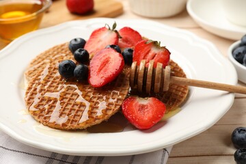 Tasty Dutch waffles (stroopwafels) with honey, strawberries and blueberries on light wooden table, closeup