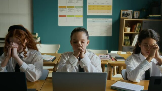 Medium shot of biracial teen girl in uniform reciting prayer sitting with classmates in front of laptops preparing for computer science lesson in modern religious school