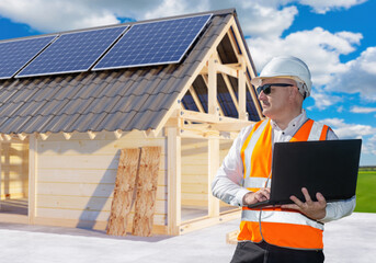 Fototapeta premium Builder near energy efficient house. Man architect holding laptop. Construction of house with solar panels on roof. Development of energy efficient housing. Man designer of eco cottage