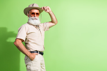 Elderly man in safari clothing with hat and glasses standing against green background in stylish...
