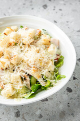 Fresh Caesar salad with romaine lettuce, grilled chicken, croutons, Parmesan cheese, and creamy dressing served in a white bowl on a gray speckled surface, top view.