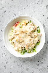 Fresh Caesar salad with romaine lettuce, grilled chicken, croutons, Parmesan cheese, and creamy dressing served in a white bowl on a gray speckled surface, top view.
