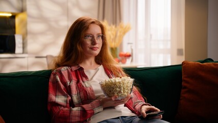 Young Caucasian woman teen girl watching TV changing channels television films show amazed say wow relaxing at home holding bowl with popcorn choosing movie weekend in living room resting on couch