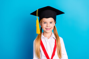 Portrait of cheerful young girl in graduation cap with red ribbon on blue background, symbolizing...