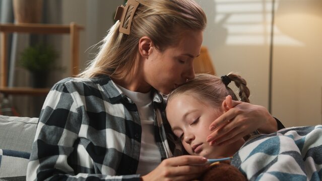 Family at home mother caring daughter kid girl measure temperature with thermometer worried about fever woman mum kissing child forehead cuddling hugging health medicine problem symptoms illness sick