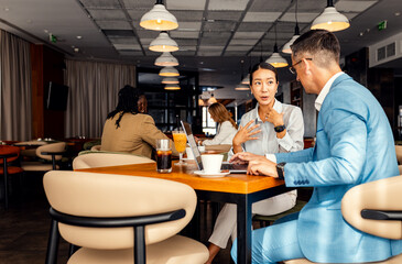 Asian woman and coworker talking about work while using laptop in modern coffee shop.