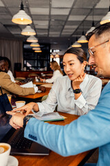 Asian woman and coworker talking about work while using laptop in modern coffee shop.