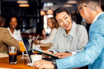 Asian woman and coworker talking about work while using laptop in modern coffee shop.