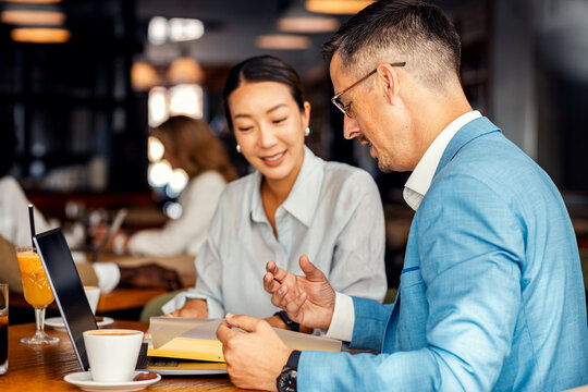 Asian woman and coworker talking about work while using laptop in modern coffee shop.