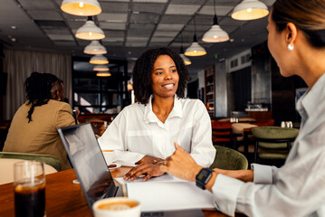 Hispanic woman and coworker talking about work while using laptop in modern coffee shop.