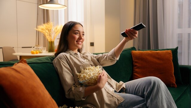 Caucasian woman girl falling on comfortable couch with bowl of popcorn turning on TV laughing enjoying snack watching television movie show cozy at home entertainment relaxing happy leisure weekend