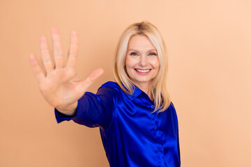 Smiling blonde woman in blue blouse gesturing with hand forward in stylish pose against solid beige...