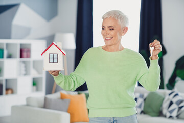 Smiling mature woman holding a model house and key in a cozy living room, symbolizing real estate...