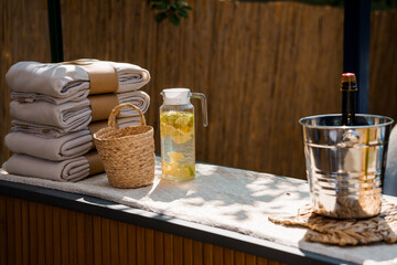 Luxurious Outdoor Relaxation Scene with Rolled Towels, Lemon Water, Wine Bottle, and Metal Bucket on Wooden Patio Table.
