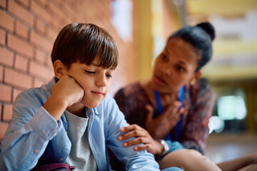 Sad schoolboy with supportive teacher in school hallway.