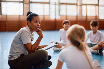 Black female coach and group of kids talking on physical education class at school gym.