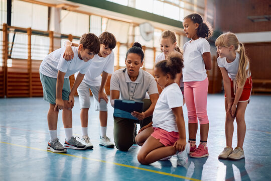 Black sports teacher and her students going through exercise plans at school gym.