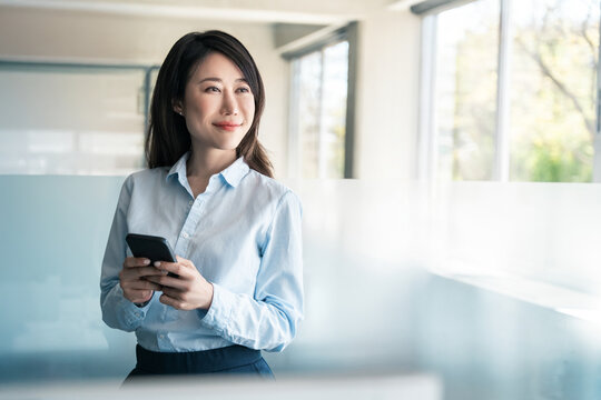 Asian Korean business woman manager using cell phone mobile trade app standing in office. Middle age female ceo businesswoman working on smartphone cellphone application looking dreaming at copy space