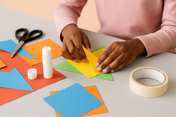 Child crafting colorful geometric shapes with paper, glue, and scissors at a table for creative arts and crafts project