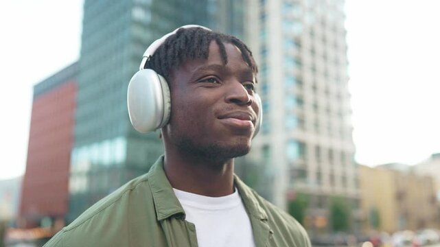 Relaxed African American young man wearing wireless headphones outdoors. Male with closed eyes enjoying music while standing near modern city buildings. Peaceful moment in urban environment. Close up.