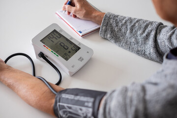An elderly woman using a medical device to measure blood pressure and taking notes on the results -...