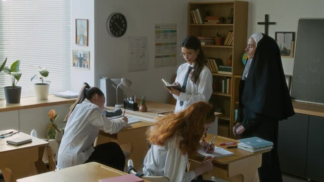 Teen Caucasian student girl in uniform reading Bible in front of class under supervision of senior nun teacher with her classmates writing in notebooks studying in modern religious school