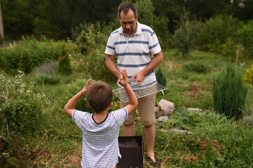 Father and son grilling sausages on fire in backyard garden. Family time, involved parenting,...