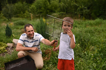Father and son grilling sausages on fire in backyard garden. Family time, involved parenting,...