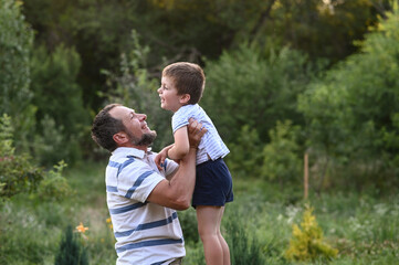 Fototapeta premium Happy father and son playing outside in garden in summer in sun. dad roll son on neck, roll on shoulders, play throws up. involved parenting, time with children. leisure without gadgets. Father's day