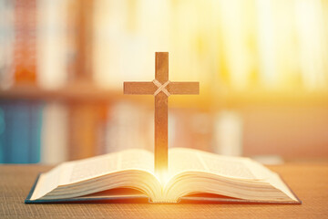 Wooden Christian cross standing on open Bible with glowing sunlight background