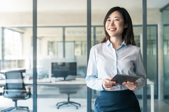 Smiling businesswoman CEO holding digital tablet using fintech tab online for work standing in modern office. Dreaming thoughtful portrait of mature Asian woman business female manager. Copy space