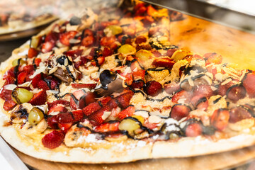 Colorful fruit dessert pizza behind glass. Sweet toppings, gourmet snack, and indulgent street food with fresh strawberries, banana slices, and melted chocolate.