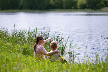 Mother and son sitting by the lake on a summer day, mother pointing into the distance, bonding in...