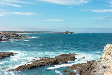 Fototapeta premium View of the rocky coastline near Ilha do Pessegueiro, Portugal. Blue Atlantic waters crash against the rugged shore under a bright sky. Perfect seascape for travel and nature themes.