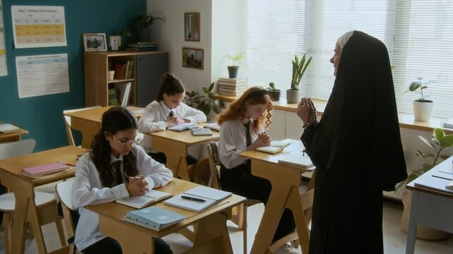 Medium shot of mature Caucasian nun in veil with rosary beads in hand praying with teenage students sitting at school desks in light modern classroom during Bible class in religious school