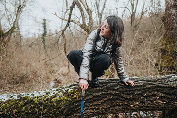 A woman wearing outdoor clothing smiles while crouching on a big mossy log in a snowy forest,...