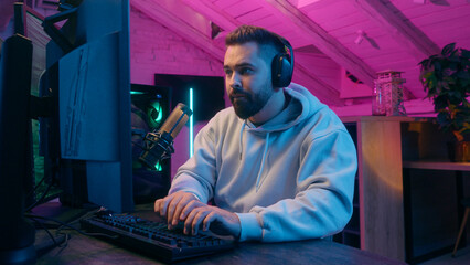 Fototapeta premium Concentrated guy male gamer streamer man wearing headphones gaming at computer focused streaming in neon light room indoors online match modern digital entertainment tech internet game playing addict