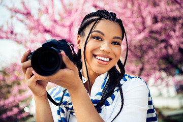 Cheerful Young Woman Holding Camera Outdoors with Beautiful Blooming Trees in Background on a Sunny...