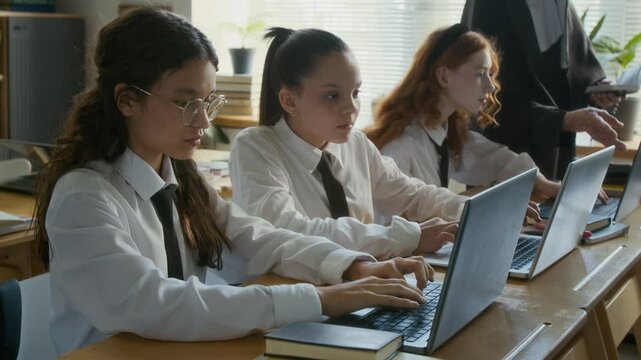 Medium shot of concentrated multiethnic teen girls working on laptops during computer science lesson in religious school with supervising nun teacher helping them