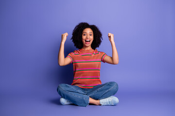 Joyful young woman in casual clothing celebrating success while seated against a vivid purple...