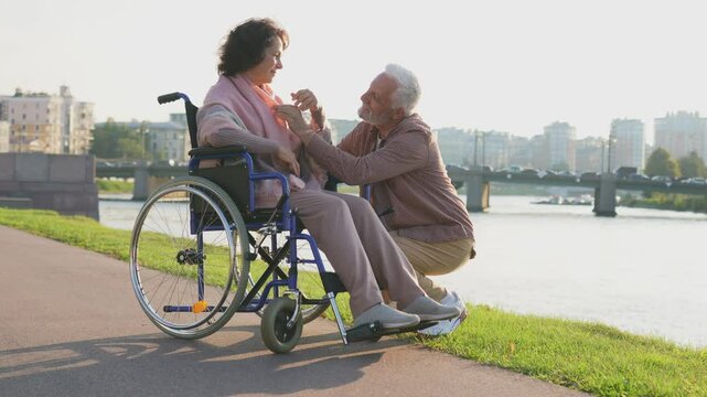 Senior woman in wheelchair walking with caregiver old man on road. Elderly family couple man supporting taking care of paralyzed woman in chair for people with disability outdoor. Rehabilitation