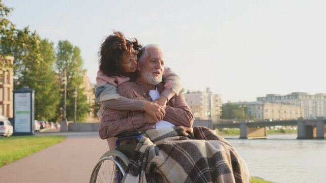 Old man in wheelchair walking with caregiver senior woman on road in park. Elderly family couple woman supporting embracing paralyzed man in chair for people with disability outdoor. Rehabilitation