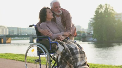 Senior woman in wheelchair walking with caregiver old man on road in park. Elderly family couple man supporting embracing paralyzed woman in chair for people with disability outdoor. Rehabilitation - Powered by Adobe