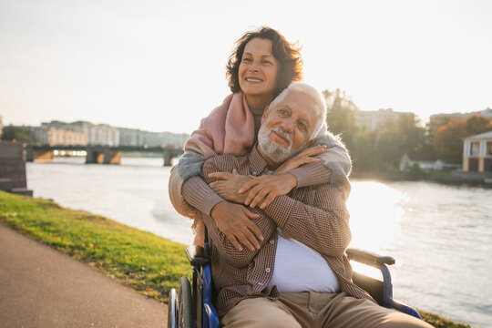 Old man in wheelchair walking with caregiver senior woman on road in park. Elderly family couple woman supporting embracing paralyzed man in chair for people with disability outdoor. Rehabilitation