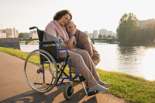 Senior woman in wheelchair walking with caregiver old man on road. Elderly family couple man supporting taking care of paralyzed woman in chair for people with disability outdoor. Rehabilitation