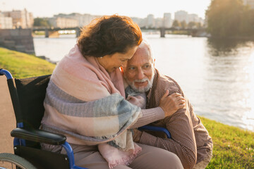 Senior woman in wheelchair walking with caregiver old man on road in park. Elderly family couple man supporting embracing woman in chair for people with disability outdoor. Rehabilitation